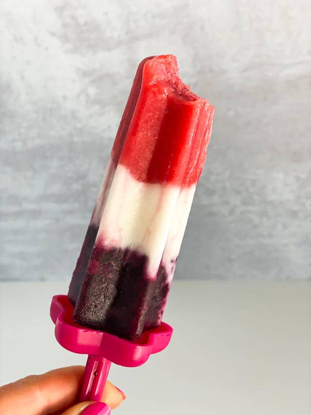 bite taken out of red, white and blue popsicle, held in wmen's hand against grey background