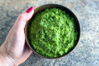 horizontal image of vegan low FODMAP kale pesto in a brown bowl, held by woman's manicured hand over stove surface