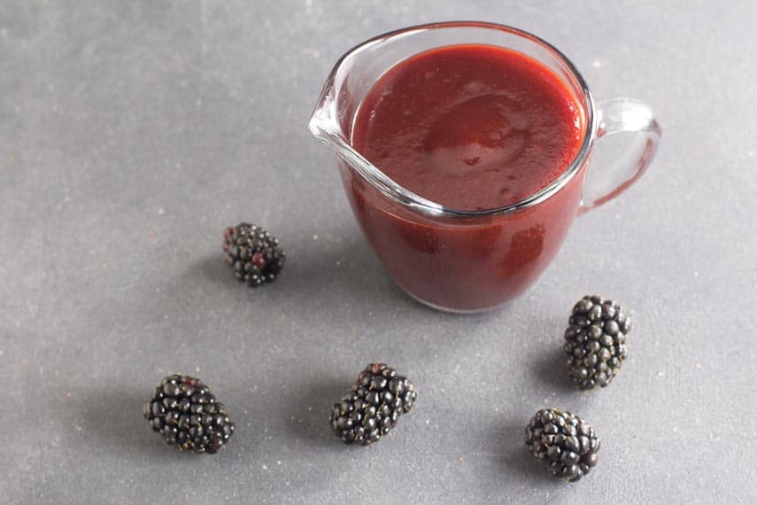 low FODMAP Blackberry Maple BBQ Sauce in clear pitcher, dark table; blackberries scattered about