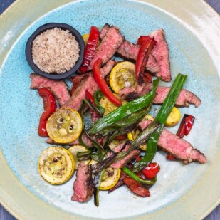 Low FODMAP Grilled Rib Eye with Smoked Salt and Charred Vegetables on ceramic plate against a blue background; overhead view