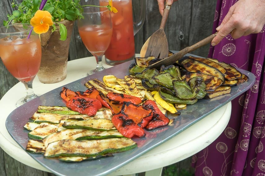 low FODMAP grilled vegetables on a platter with a woman servings them with wooden servers; iced tea in background