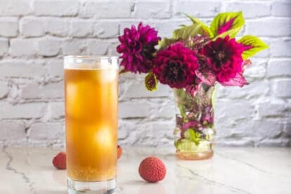 Low FODMAP Iced Black tea with Lychees in tall clear glass; white background and magenta flowers in a small vase