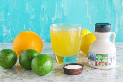 Maple Lime Low FODMAP Sports drink in a glass surrounded by ingredients against an aqua backdrop_
