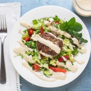 overhead shot of low FODMAP Lamb Burgers with Tahini Sauce on salad and white plate; linen napkin and fork alongside