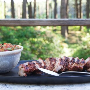 Quick Grilled Pork Tenderloin sliced on a black platter on the deck with grilled tomato peach salsa alongside