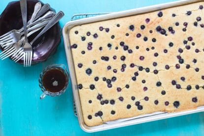 Low FODMAP Sheet Pan Pancakes with Blueberries in pan on cooling rack against aqua background; maple syrup on the side in glass pitcher