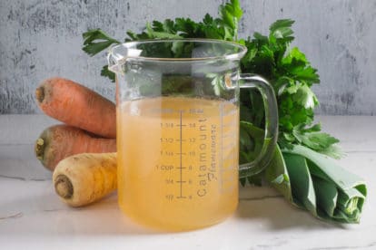 A glass measuring container filled with homemade Low FODMAP vegetable broth, in front of fresh carrots, parsnips, parsley and leeks.