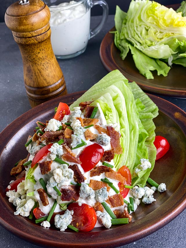 vertical image of Loaded Low FODMAP Wedge Salad sprinkled with bacon, blue cheese, tomatoes and chives on brown ceramic plate; peppermill in background